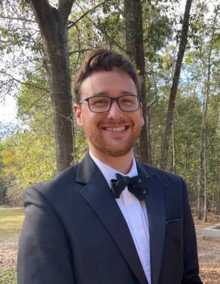 Cam Chatham stands outside near a grove of trees, wearing glasses and a bowtie, smiling toward the camera. 