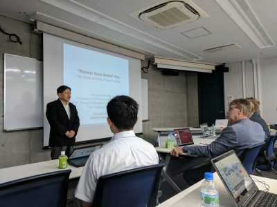 A lecturer stands in front of his slides while others listen.