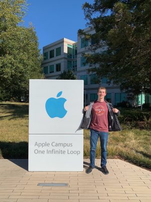 A young man wearing a black coat and maroon Hokies shirt stands next to a sign saying Apple Campus One Infinite Loop and the Apple logo . It is a sunny day with blue skies and green trees around.