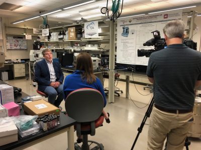 Tim Long is interviewed by an anchor from W-S-L-S and a cameraman in the M-M-D-C lab at Virginia Tech. Long wears a navy blazer and white button down shirt.