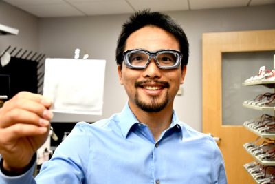 Associate Professor Feng Lin, wearing a blue dress shirt and goggles, holds a pouch battery cell in his battery-testing lab at Davidson Hall. White in color, square in shape, the battery has two prongs sticking out of one side. Behind Feng is a rack with dozens of lithium-ion batteries being tested in charge cycles. Photo for Virginia Tech by Steven Mackay. 
