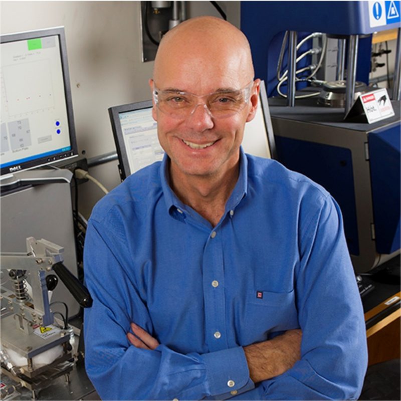 A man wearing a blue shirt and safety glasses smiles while standing in front of laboratory equipment.