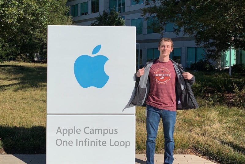 A young man wearing a black coat and maroon Hokies shirt stands next to a sign saying Apple Campus One Infinite Loop and the Apple logo . It is a sunny day with blue skies and green trees around.