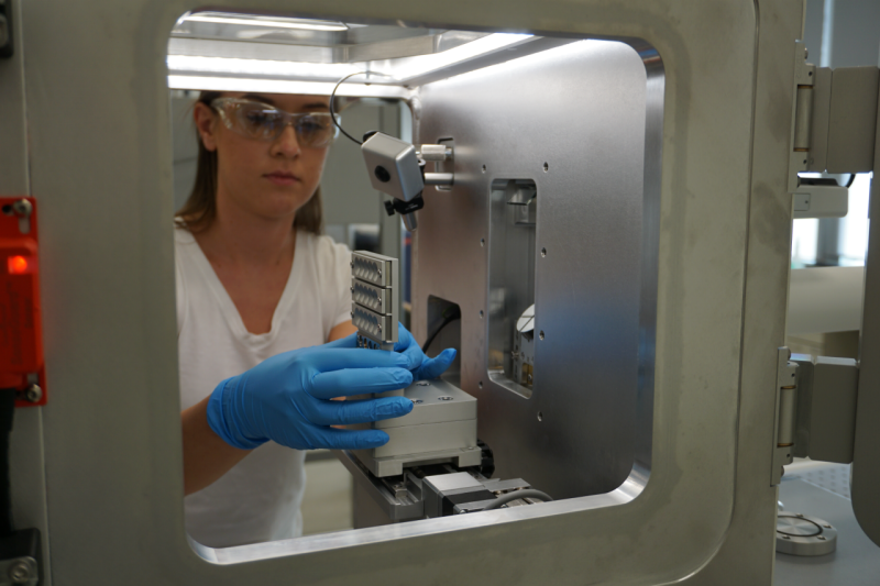 A woman wearing safety glasses is seen through the window of a large laboratory instrument.